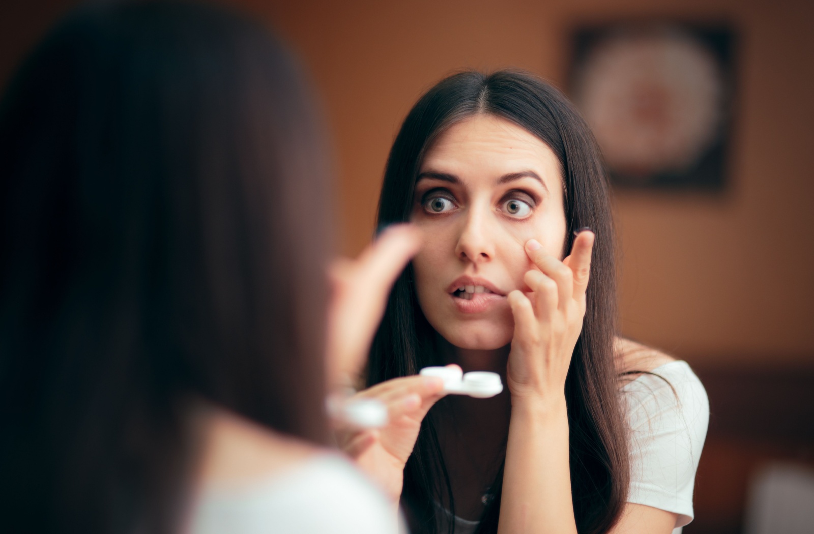 A young adult looks in the mirror while holding their contact lens case, worried the lenses are irritating their eyes